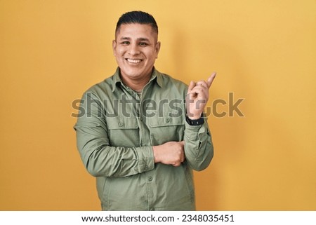 Hispanic young man standing over yellow background with a big smile on face, pointing with hand finger to the side looking at the camera. 