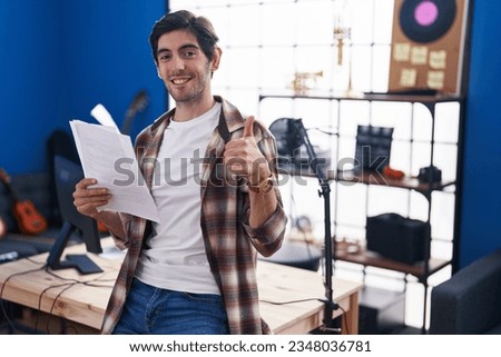 Young hispanic man reading music sheet at music studio smiling happy and positive, thumb up doing excellent and approval sign 