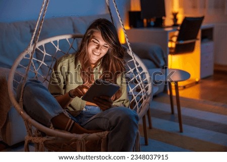 Beautiful young woman sitting in hanging chair surfing the net using tablet computer while relaxing at home Royalty-Free Stock Photo #2348037915