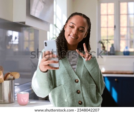 Teenage Girl At Home In Kitchen Posing For Selfie To Upload To Social Media On Mobile Phone
