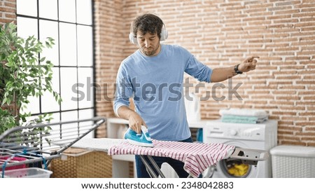 Young hispanic man listening to music ironing clothes dancing at laundry room