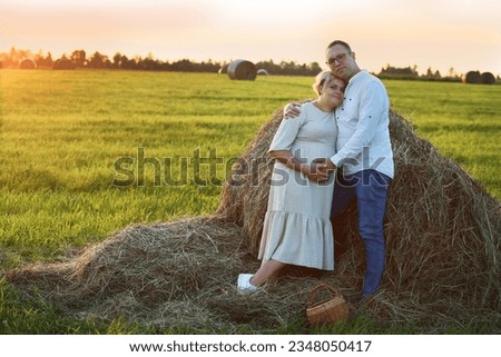 blond pregnant woman in white dress and her husband with basket on hay summer field background