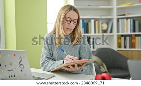 Young blonde woman student using laptop writing on notes at library university
