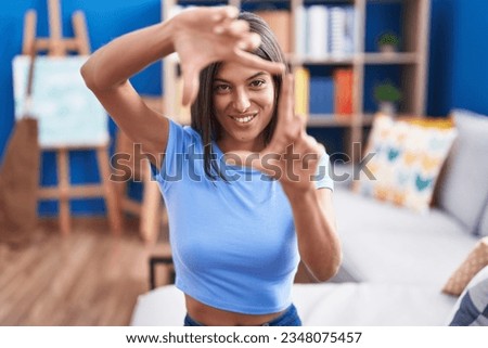 Brunette young woman sitting on the sofa at home smiling making frame with hands and fingers with happy face. creativity and photography concept. 