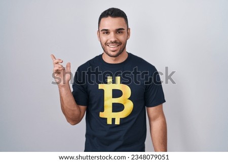 Young hispanic man wearing bitcoin t shirt cheerful with a smile on face pointing with hand and finger up to the side with happy and natural expression 