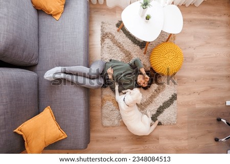 Top view of beautiful young woman relaxing at home and spending time with her pet, Labrador dog, both lying on the living room floor, woman petting dog Royalty-Free Stock Photo #2348084513
