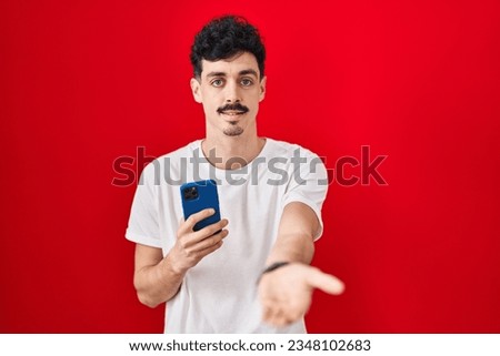 Hispanic man using smartphone over red background smiling cheerful offering palm hand giving assistance and acceptance. 