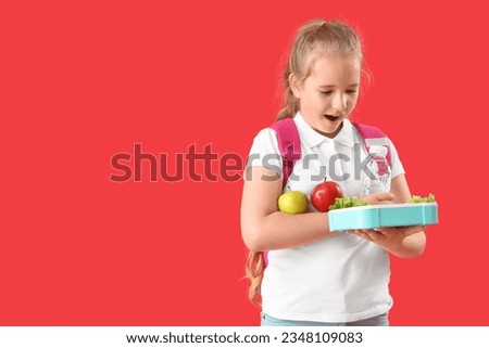 Surprised little girl with backpack, lunchbox and apples on red background