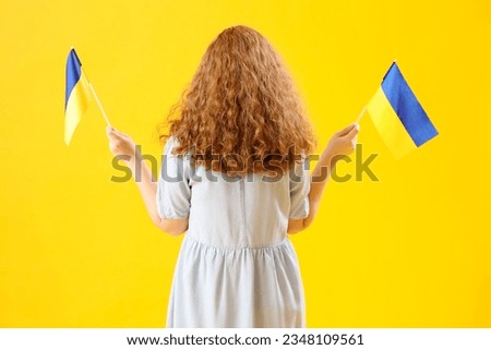 Beautiful young woman with Ukrainian flags on yellow background, back view