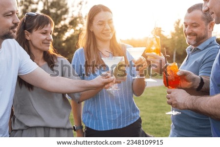 Meeting friends in the backyard of the house or in open air cafe. Young men and women are holding glasses with alcoholic beverages outdoors. Smiling people clink glasses with cocktails and make toast.