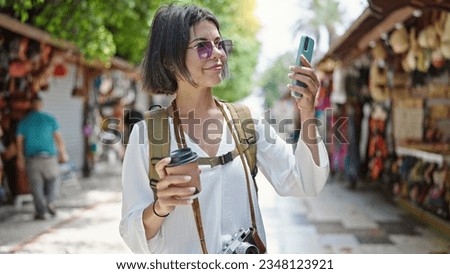 Young beautiful hispanic woman tourist smiling confident using smartphone drinking coffee at street market