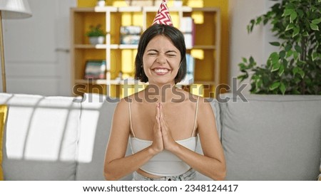 Young beautiful hispanic woman wearing birthday hat sitting on sofa praying at home