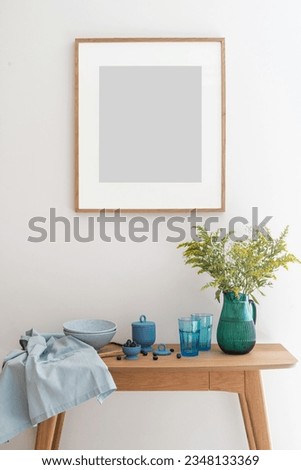 front view on wall picture in frame, wooden table with flowers in vase, clean glasses and plates, ceramic food container with lid and fresh blueberries