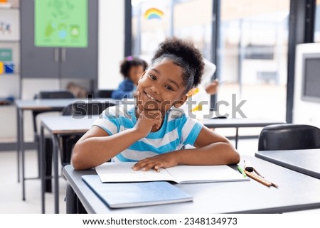 Portrait of happy african american schoolgirl sitting at desk in school classroom. Education, childhood, elementary school and learning concept. Royalty-Free Stock Photo #2348134973