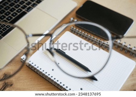 View through glasses on working wooden table with notepads, pen, smartphone and open laptop