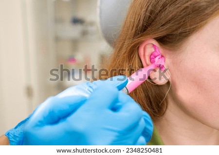 A doctor fills a patient's ear canal with a silicone material, pink, made using a syringe for making earplugs in a hearing clinic. Production of inserts to order Royalty-Free Stock Photo #2348250481