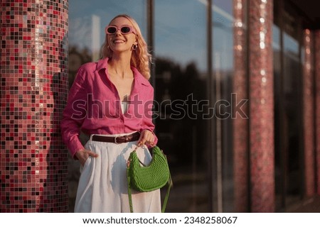 Fashionable happy smiling blonde woman wearing trendy summer outfit with pink sunglasses, linen shirt, belt, white shorts, holding small green faux leather bag, posing in street of city. Copy space Royalty-Free Stock Photo #2348258067