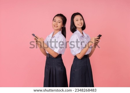 Two teenage Asian female students with short hair and long hair, smiling cute, wearing school uniforms, using two smartphones, standing back to back. Photo shoot in pink background studio