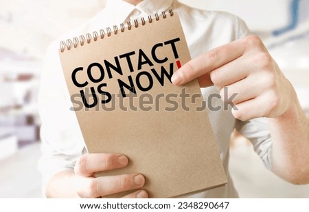 Text CONTACT US NOW on brown paper notepad in businessman hands in office. Business concept
