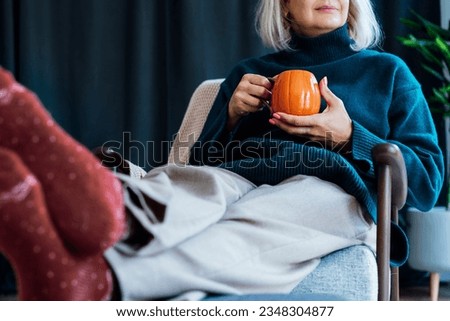 Cropped mature middle aged woman in sweater and warm socks relaxing in armchair with pumpkin shaped cup of hot coffee or tea drink. Cozy calm autumn holidays at home. Fall hygge mood concept. Royalty-Free Stock Photo #2348304877