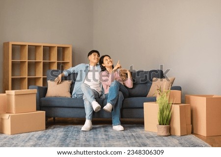 Great Real Estate Offer. Happy Japanese Young Spouses After Moving Sitting On Sofa Among Cardboard Boxes In Living Room, Woman Pointing Finger Aside At Wall Dreaming About Renovation At New Home Royalty-Free Stock Photo #2348306309