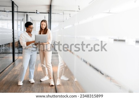 Businesswoman with laptop explaining project details to colleague at office hallway. Business partners having meeting in coworking office space. Two female diverse employees, full length