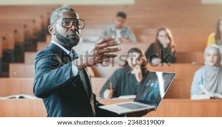 African American male professor standing in front of students in college with laptop and talking to them while having lection. High school. Man teacher explaining subject with computer. Close up. Royalty-Free Stock Photo #2348319009