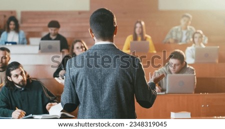 Rear on young Caucasian male professor speaking at lection at University.Back view. Lecturer at high school. Students on background. Royalty-Free Stock Photo #2348319045