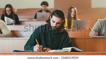 Portrait of happy handsome Caucasian male student sitting in auditorium, reading textbook, doing marks and smiling to camera. Guy read book and marking something interesting at lection. Study. Royalty-Free Stock Photo #2348319059