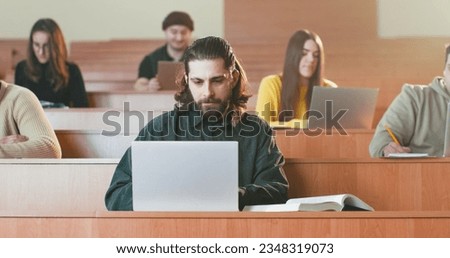 Young handsome Caucasian male student sitting at desk in auditorium at laptop and reading textbook. Education concept. Guy read book and using computer at lection in college. Obtaining master degree. Royalty-Free Stock Photo #2348319073
