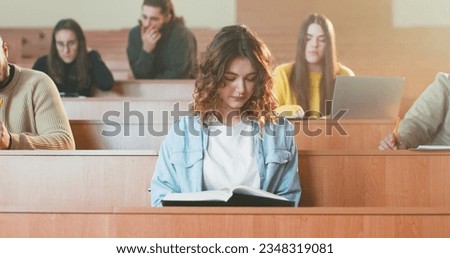 Close up of young pretty Caucasian female student sitting at desk in auditorium and reading textbook. College education concept. Girl read book at lection in University. Obtaining knowledge. Royalty-Free Stock Photo #2348319081