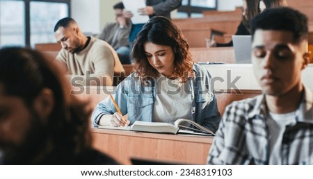 Young pretty Caucasian female student listening lection at college, writing down and using textbook. Reading book at seminar at high school. University study concept. Studying in classroom. Royalty-Free Stock Photo #2348319103