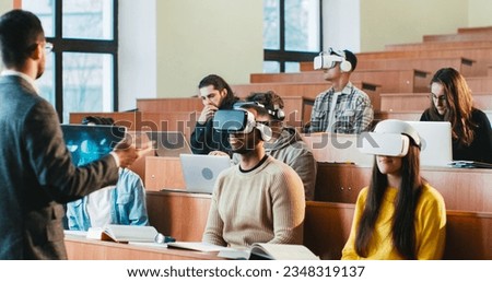 Rear of professor having speech in front of mixed-races students in VR glasses and laptops. Multiethnic males and females studying in technologic high school and listening to teacher at seminar. Royalty-Free Stock Photo #2348319137