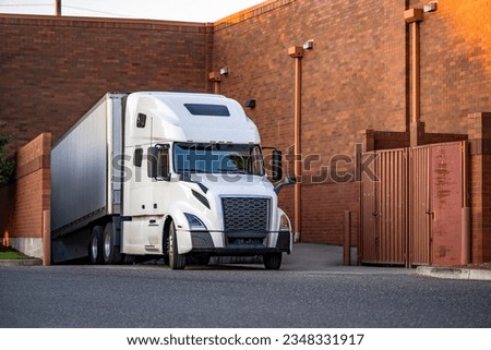 Bonnet powerful industrial big rig semi truck tractor unloading delivered in dry van semi trailer commercial cargo standing in narrow bricks store cargo dock at twilight time Royalty-Free Stock Photo #2348331917