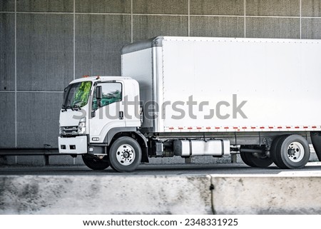 Industrial standard clean white day cab middle rig cab over semi truck tractor transporting commercial cargo in box trailer driving on the divided highway road under the bridge in urban city limit Royalty-Free Stock Photo #2348331925
