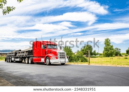 Long hauler big rig red semi truck tractor with grille guard and loaded by pipes flat bed semi trailer standing on the local road take a break for truck driver rest according to log book schedule Royalty-Free Stock Photo #2348331951