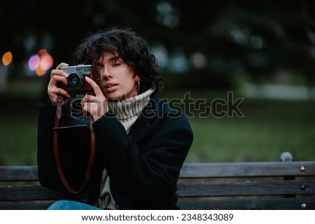 Cropped shot of a pretty caucasian dark haired curly girl sitting on a bench at the park and taking photos on her old retro camera. Enjoying a beautiful winter day outside in a nature. Copy space.
