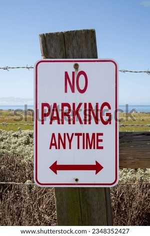 No parking anytime sign on the fence with barbed wire. No parking any time sign with ocean coast at background. No parking anytime sign outdoor. Vertical arrangement