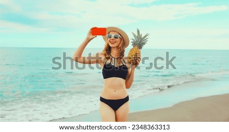 Summer vacation, happy smiling young woman holding pineapple taking selfie with smartphone wearing bikini swimsuit and straw hat on the beach on sea coast background