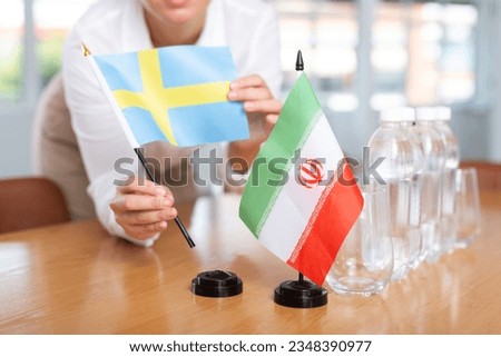 Little flag of Iran on table with bottles of water and flag of Sweden put next to it by positive young woman 