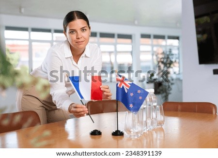Young woman in business clothes puts flags of France and New Zealand on negotiating table in office
