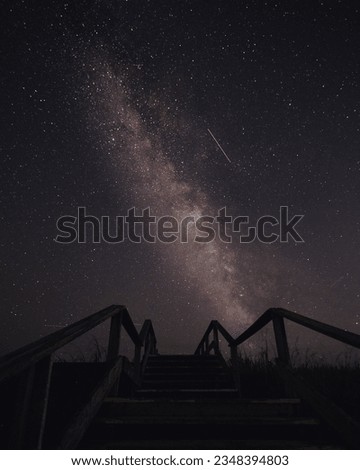 A rustic wooden staircase rises towards the moonlit beach, illuminated by the gentle glow of stars. Above, the Milky Way stretches across the sky in a mesmerizing display of cosmic beauty. 
