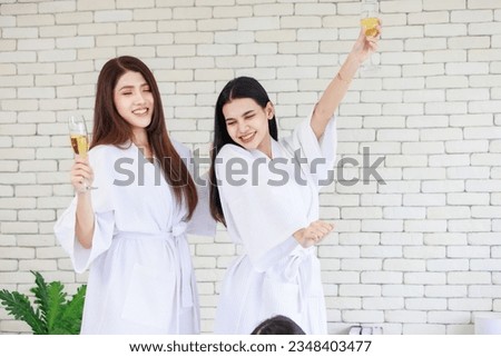 Millennial two Asian beautiful drunk girlfriends in white clean bathrobes celebrating party relaxing resting holding drinking tall glasses of golden sparkling champagne in spa massage studio resort.