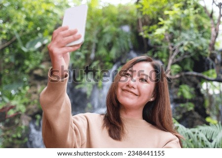 Portrait image of a beautiful young asian woman using mobile phone to take a selfie in the garden with waterfall