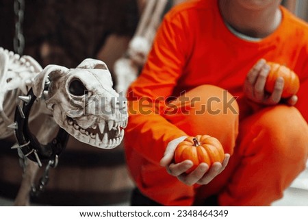 Young teen boy with painted face spider while all hallows eve photo session indoor. Stylish child in orange clothes celebrates halloween holiday near human skeleton with skull