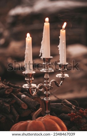 Halloween pumpkin and burning candlestick by three candles at dark background twilight in open air. All hallows eve natural decoration with fallen leaves and branch in windy weather at autumn season