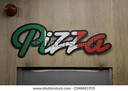 The written word Pizza restaurant sign in the colours of the Italian Flag, Green, White and Red with a black outline, on a wooden Wall over a grey metal door. Theres a red warning light above the sign Royalty-Free Stock Photo #2348481935