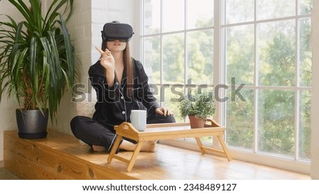 Beautiful young girl in a virtual reality headset. Drinking morning coffee and putting on VR goggles. Happy cute woman touching something using modern 3d vr glasses indoors.