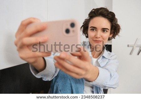 Stylish young girl blogger, takes selfie in her kitchen, poses for photo on mobile phone, using camera app for a video with filters.