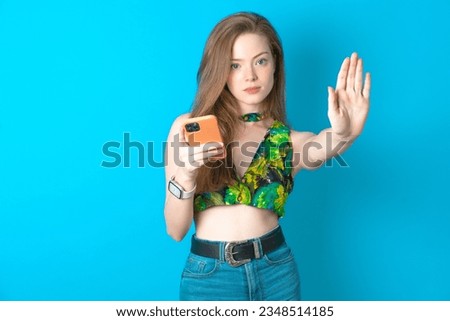 young beautiful blonde woman wearing green top standing over blue studio background using and texting with smartphone with open hand doing stop sign with serious and confident expression.